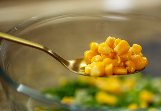 Extreme close-up of shiny yellow corn kernels piled on a golden spoon held over a glass bowl of fresh green salad - Powered by Adobe