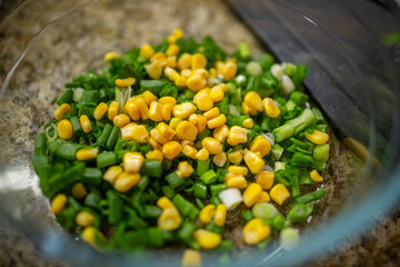 Close-up of vibrant yellow corn kernels mixed with freshly chopped green onions inside a clear glass bowl on a granite countertop