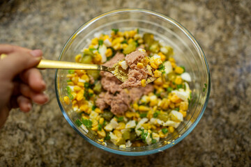 Close-up of canned tuna being added and mixed into a vibrant glass bowl of salad with corn, eggs, and chopped pickles