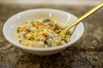 Close-up of a white bowl of creamy tuna salad being served or eaten with a golden spoon on a brown granite kitchen countertop