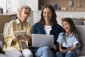 Laughing granny mom daughter sitting on sofa using diverse gadgets