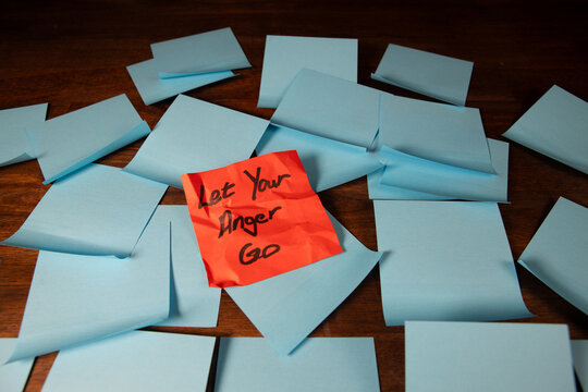 Red crumpled sticky note on a pile of blue sticky notes placed on a dark wood surface with text reading let your anger go.