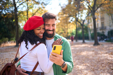 Happy interracial couple hugging and looking at a smartphone in an autumn park. Close-up travel portrait in natural light that conveys love using mobile, connection and joyful lifestyle.