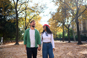 Interracial couple walking through an autumn park, smiling and holding hands. Casual travel portrait in natural light with leaves and trees, conveying romance, connection and joyful lifestyle.