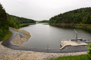 A calm reservoir lies amidst lush forest with a large bridge visible in the distance. Scenic natural landscape with water, trees, and peaceful outdoor environment.