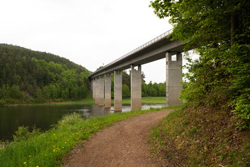 A massive bridge stretches across a river with forested hills and mountains in the background. Scenic natural landscape, calm water, and tranquil outdoor environment.