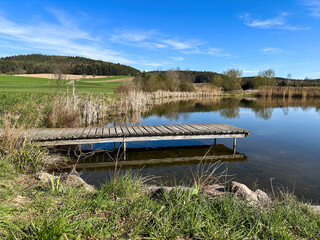 A small wooden dock stretches into a peaceful lake, used for small boats, fishing, or quiet relaxation. Serene water, natural surroundings and soft light create a tranquil outdoor scene.