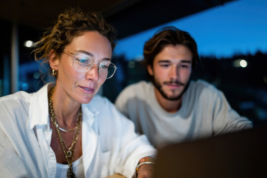A woman and a man collaborate over a laptop in dim evening light, showcasing teamwork, focus, and the modern workplace in a cozy environment.