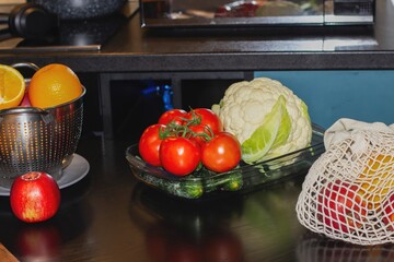 Fresh cucumbers and tomatoes laid out on a plate on the kitchen table, with fresh fruits and vegetables in the background. Useful ingredients for making salad at home