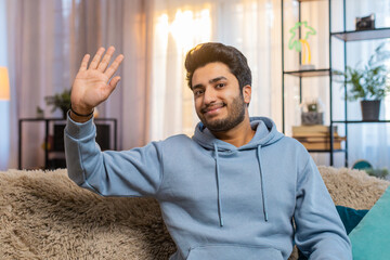 Portrait of cheerful Indian young man smiling at camera, waving, gesturing hello, hi, or goodbye. Happy Hispanic guy making video conference call with friend, enjoying conversation from home room.