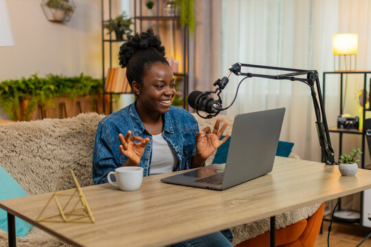 African American young woman sitting at table on home sofa records a personal podcast vlog using a studio microphone gesturing confidently. Black girl smiles warmly and enjoys sharing her voice online