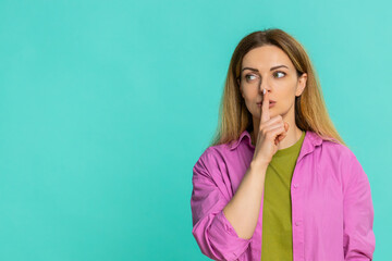 Middle-aged woman gently places finger on lips, signaling to be quiet, keep secret or avoid noise with soft serious expression. Girl isolated on blue background shows silent gesture, confidentiality.