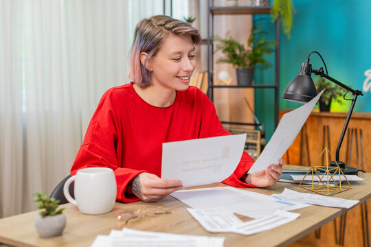 Young satisfied woman happily reviewing financial documents sitting at home table, smiling . Caucasian girl positively excited about good financial results, demonstrating successful money management.