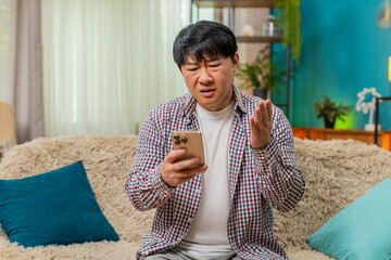 Asian man sitting on home sofa reads bad fail news on smartphone, frowning, showing disappointed frustrated mood. Chinese guy reveals negative reaction, sighs with visible displeasure and tension.