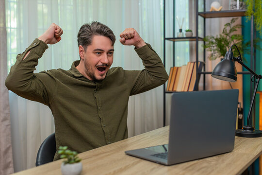 Middle-aged man at home reads laptop news good message smiles celebrating victory feeling big win success. Young adult guy working at table happy joyful after positive update enjoying accomplishment