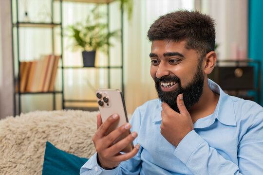 Indian man at home gets amazing smartphone message eyes widen shouts wow celebrates victory pure joy. Arabian guy on sofa raises phone smiles triumph arms up happy cheer winning great online success.