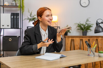 Young businesswoman at home office on smartphone speaker writes notes closing deal. Freelancer girl at table smiles after call celebrating sealed new contract and promising big growth.