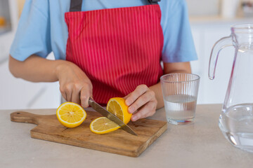 Young woman at home in kitchen cuts lemon on wooden cutting board with knife near jug of water. Hands in apron focus on clean food prep citrus slice for water drink healthy habit calm mood routine