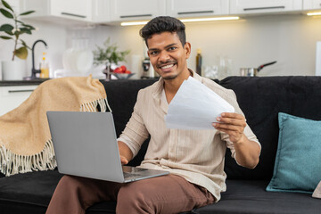 Indian man at home enters bills in spreadsheet on laptop checks papers and uses calculator to verify total. Arabian guy on sofa reviews receipts updates table saves file and records budget result log
