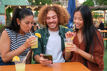 Three friends are using a smartphone and drinking cocktails at a summer festival