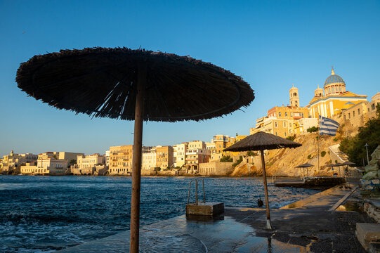 Venetian architecture of Ermoupoli and Saint Nicholas church framed by natural reed thatched parasols on a swim platform of Asteria beach on the Greek island of Syros
