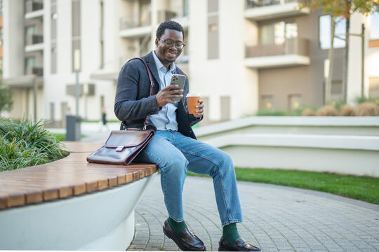 Happy young man using phone drinking coffee outdoors