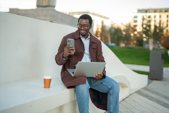Young black man multi-tasking with phone and laptop outdoors