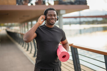 Happy man wearing headphones, carrying yoga mat by river