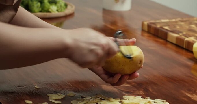 Close up of hands peeling fresh potato with peeler on wooden table, preparing ingredients for homemade vegan meal, cooking process, healthy food, rustic kitchen atmosphere, natural lifestyle