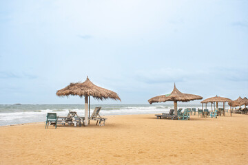 An Ivorian beach lined with thatched huts and wooden sun loungers. The Atlantic Ocean forms the horizon under a clear sky.