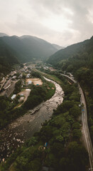 Aerial view of a river winding through a lush, mountainous valley. A road follows the river's course, passing through a small town. Overcast sky adds to the moody atmosphere.
