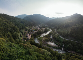 A scenic aerial view of a village nestled in a valley alongside a winding river, surrounded by lush green mountains and forests. The scene evokes a sense of tranquility and natural beauty.
