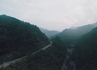 A scenic aerial view of a winding road through a lush, mountainous landscape shrouded in mist. The road cuts through dense green forests and a river valley.