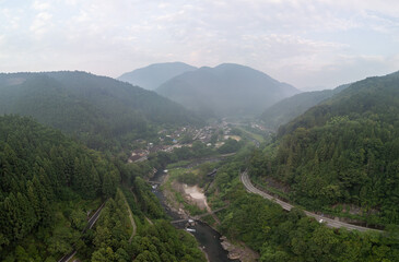 Aerial view of a lush valley with a winding river, surrounded by dense forests and mountains. A road snakes through the landscape, leading to a small town nestled among the trees. 