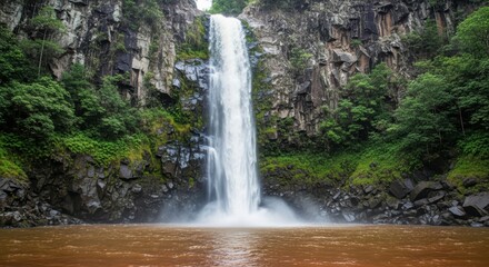 Fototapeta premium Waterfall Cascading Down Rocky Cliff into Murky Pool. Lush Green Vegetation on Cliffside Surrounding Waterfall. Dynamic Waterfall in Serene Verdant Natural Environment.