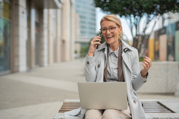 Mature woman communicating by phone and working using laptop