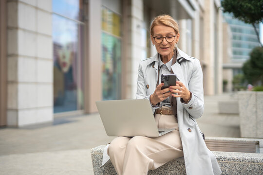 Mature businesswoman working remotely using smartphone and laptop outdoors