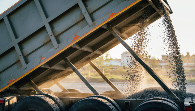 Powerful dump truck unloads gravel, creating dust clouds in warm golden hour light during construction project