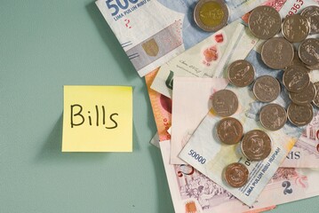A yellow sticky note with Bills written on it next to a scattered pile of banknotes and coins