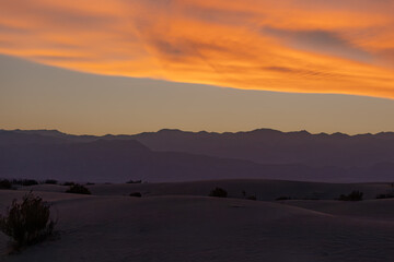 A wide desert view at sunset, with soft dunes in shadow beneath a glowing orange sky.