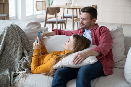 Married couple staying at home during calm weekend. Relaxed wife lying on male knees and smiling joyful husband having video call with relatives, friends. Closeness, togetherness in family relations