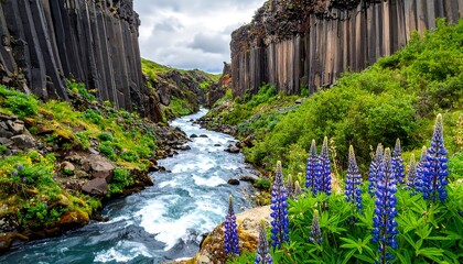 A rushing river cascades through a canyon, framed by basalt columns and vibrant green flora, under a cloudy sky. Bright purple flowers bloom