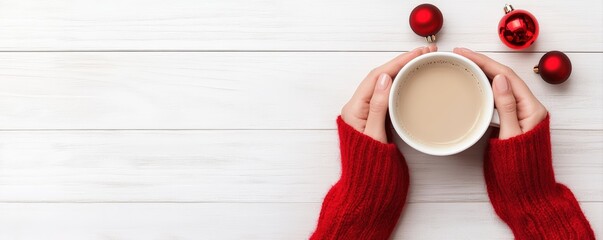 Woman's hands in a red sweater holding a hot coffee mug on a white wooden background, surrounded by Christmas ornaments
