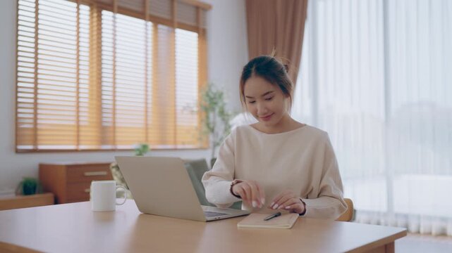Young Asian woman using laptop computer for business studying, having video call with online virtual webinar training meeting, writing notes. Focused student female working in living room at home
