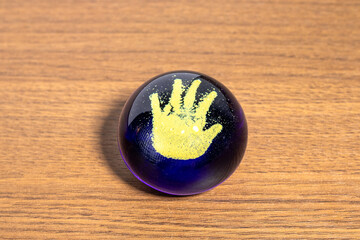A vibrant deep purple glass sphere with a textured, sparkly yellow handprint design embedded inside, resting on a warm, polished wooden table, creating a unique decorative object.