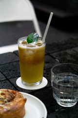 A refreshing glass of mango cold brew with ice cubes, garnished with a green leaf and served with a straw, sits beside a clear glass of water and a pastry on a dark table.