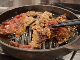 Close-up of thin slices of raw and marinated beef cooking on a round cast iron grill pan, with black chopsticks picking up a piece of meat.