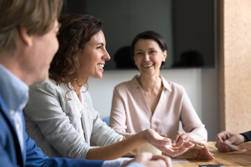 Smiling young businesswoman speak to teammates sitting around boardroom table