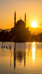 A majestic domed structure with minarets stands silhouetted against a golden sunset, reflected in tranquil water