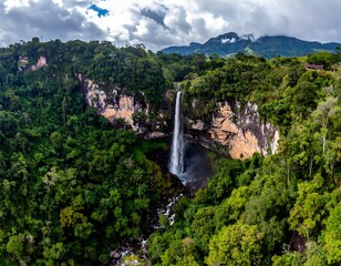 Aerial view of a majestic waterfall cascading down a cliff face surrounded by dense, vibrant green foliage. Overcast sky